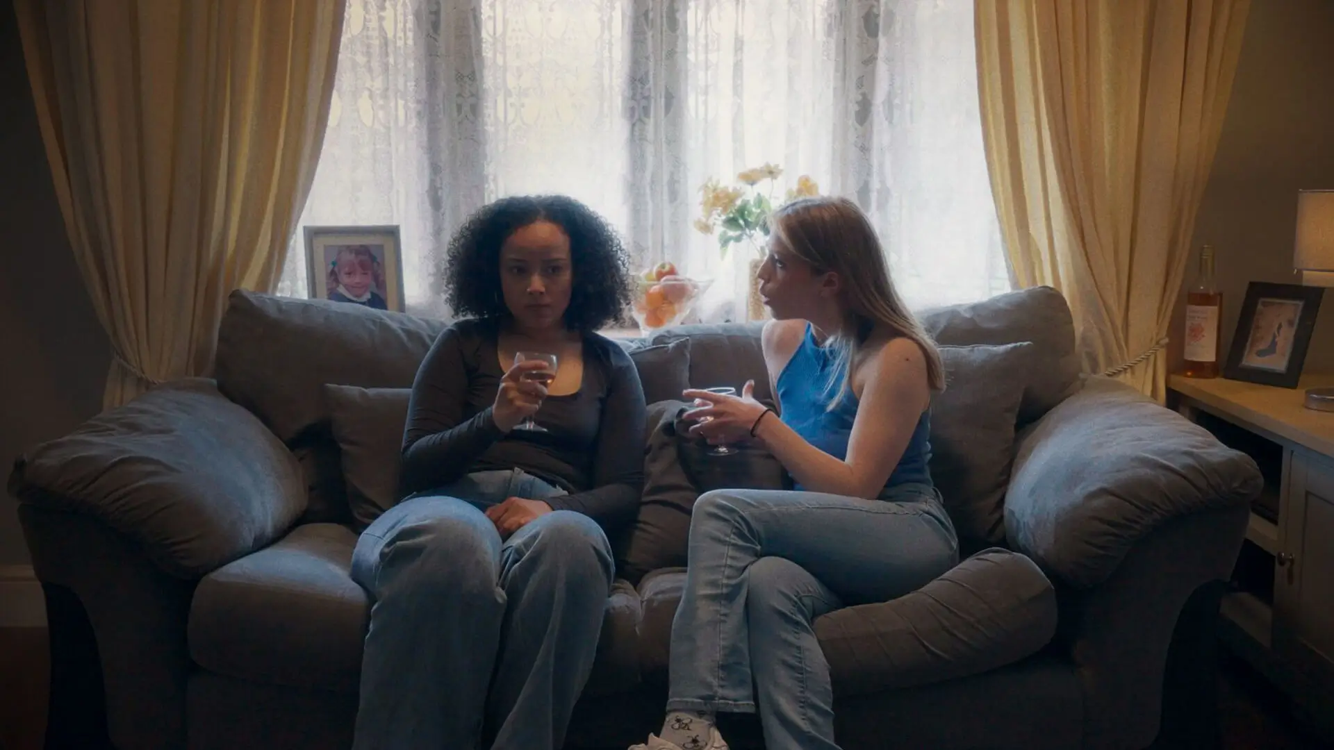 Two women, Lesley and Hannah from the film Lesley, sitting on a brown couch having a tough conversation while drinking wine. In front of a window.