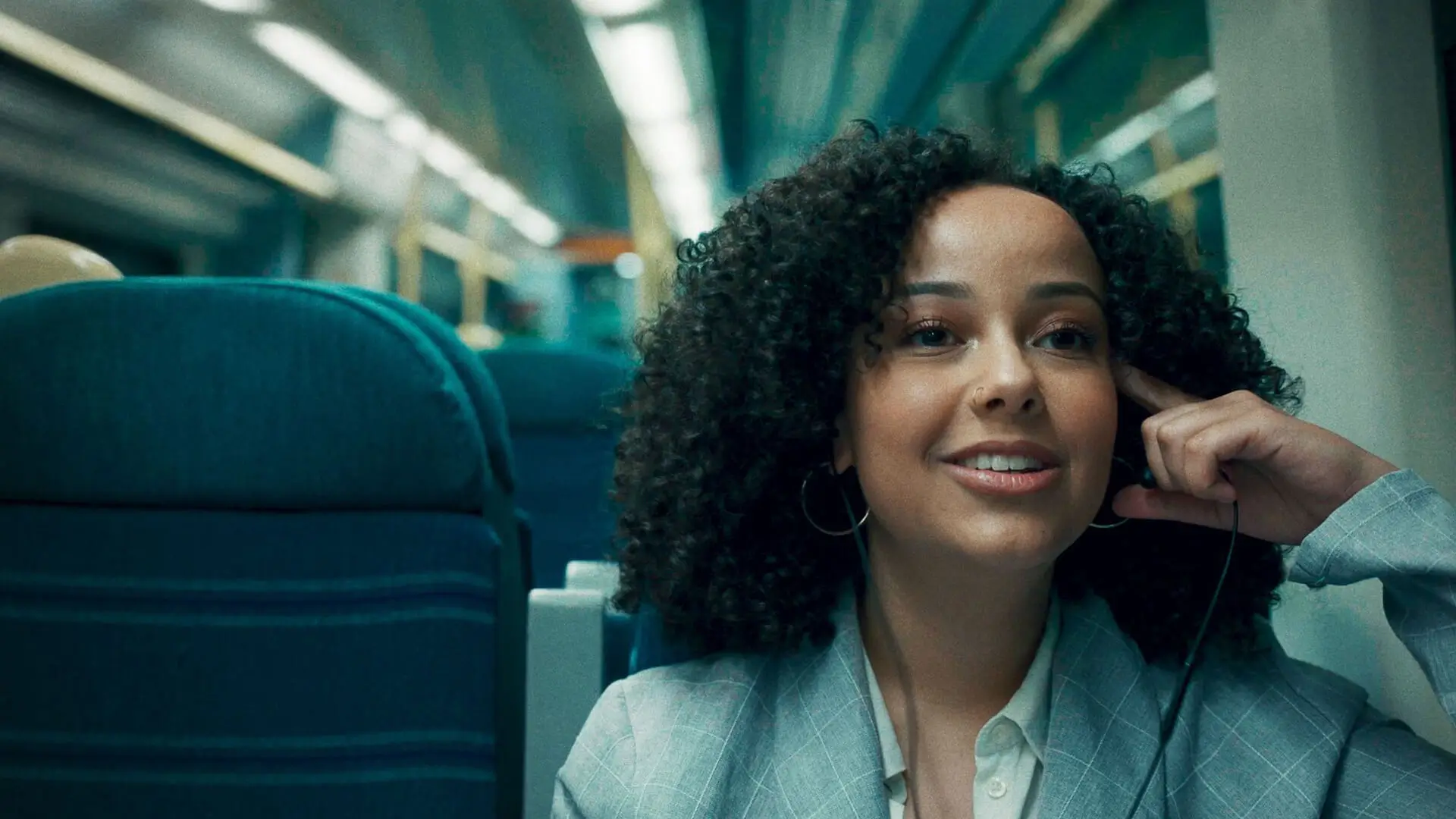 A woman, Lesley from the film Lesley, dressed smart, sitting on an empty train listening to music with one earphone in.